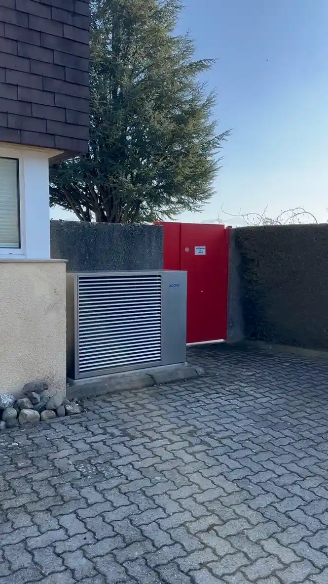 Pompe à chaleur installée pour une maison en Suisse romande Pompe à chaleur moderne installée près de l’entrée d’une maison en Suisse romande, à côté d’un portail rouge