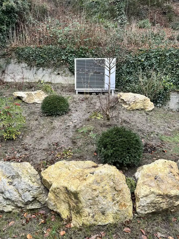Installation de pompe à chaleur discrète dans un jardin naturel en Suisse romande Pompe à chaleur installée dans un jardin paysager avec rochers et végétation en Suisse romande